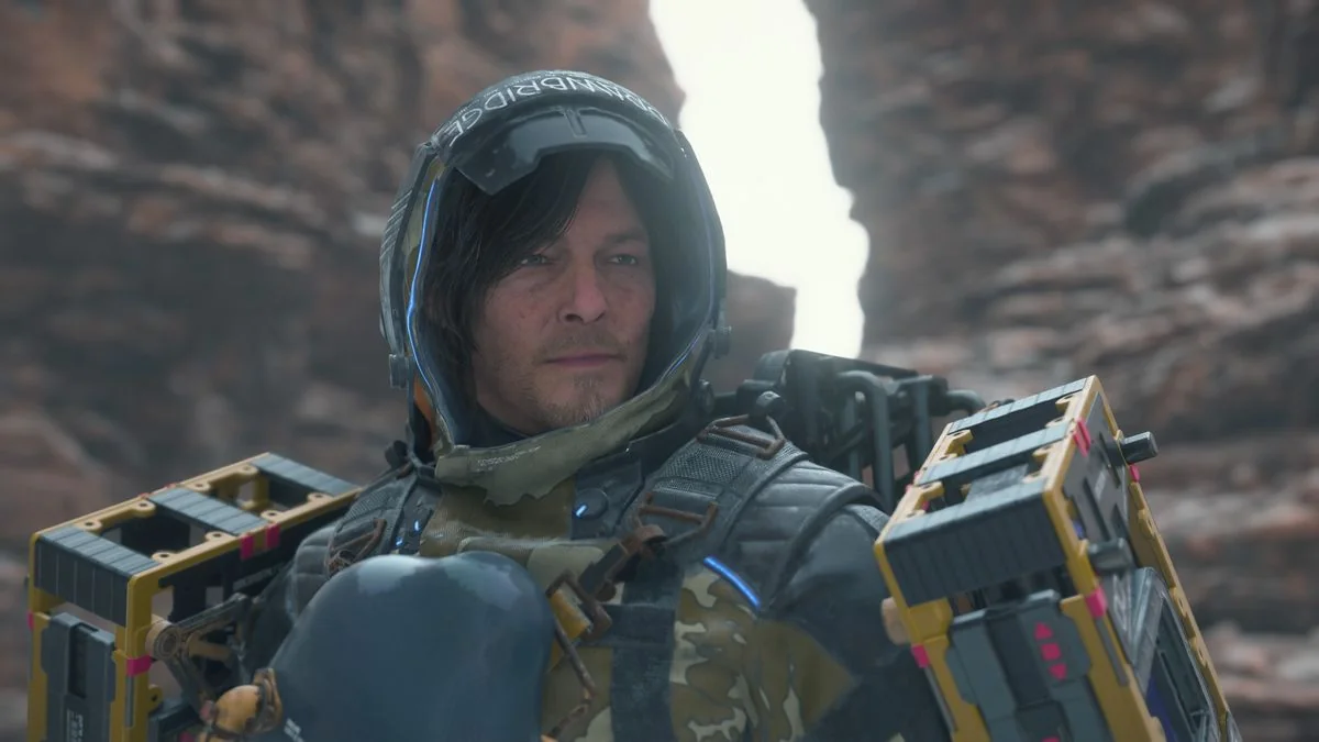 Death Stranding 2 stats: Sam mit Fracht und Mantel, schaut zur Seite, während er in einer Schlucht steht.