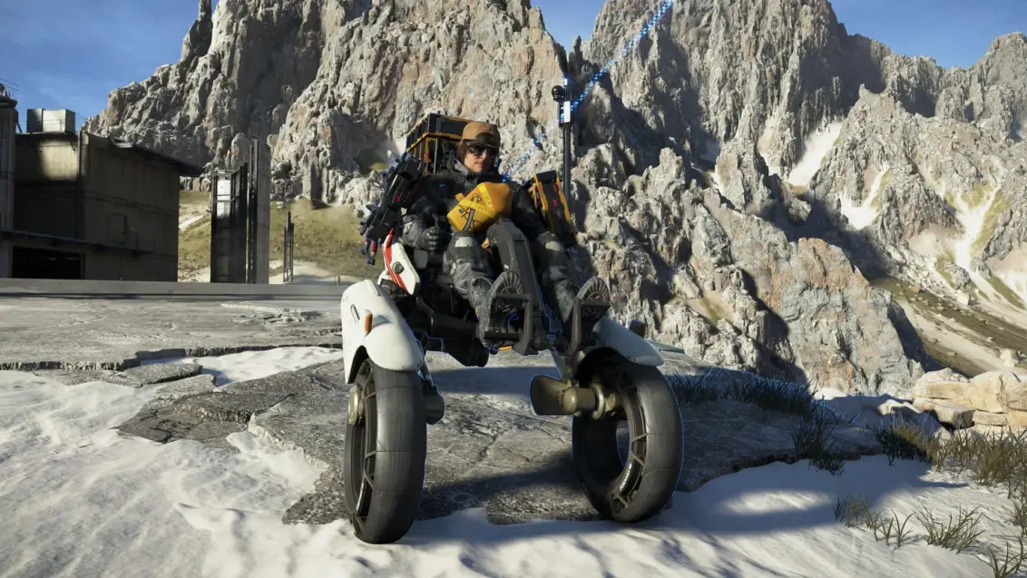 Death Stranding 2 Fahrzeuge: Eine frontale Aufnahme von Sam auf einem Tri-Cruiser, geparkt vor einer Forschungseinrichtung in einer bergigen Gegend.