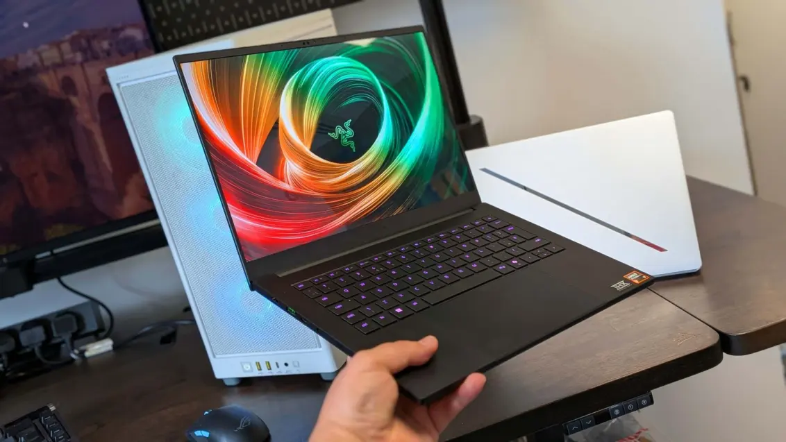 Razer Blade 14 Gaming-Laptop in einer Hand gehalten, mit einem PC und einem Asus ROG Zephyrus G14 im Hintergrund.